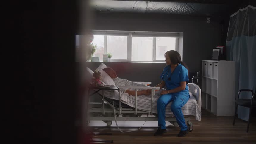 The video shows a healthcare professional in blue scrubs sitting on a hospital bed next to a patient the room is dimly lit but a large window lets in some natural light revealing the patients legs covered by a blanket and various medical equipment aro