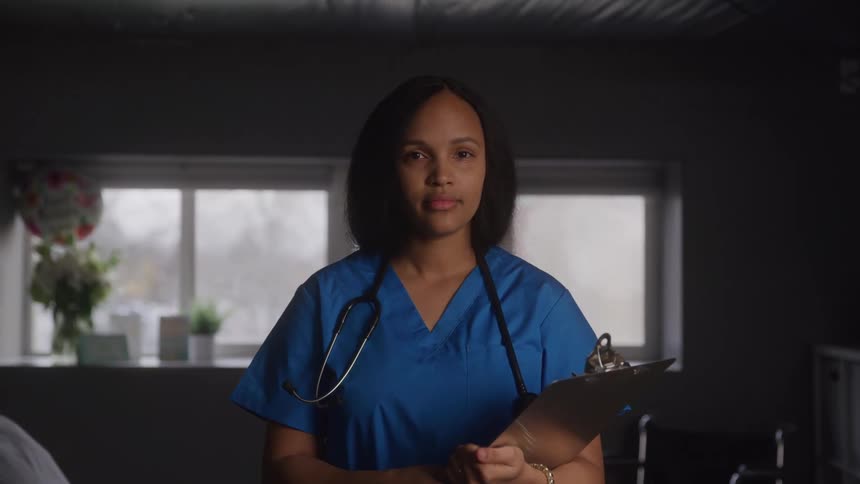 A nurse with a serious expression and a stethoscope around her neck is holding a clipboard she is wearing blue scrubs and looking directly at the camera