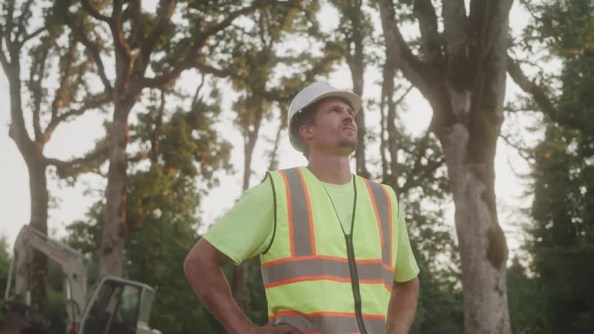 A construction worker in a safety vest and hard hat stands with hands on hips gazing upward