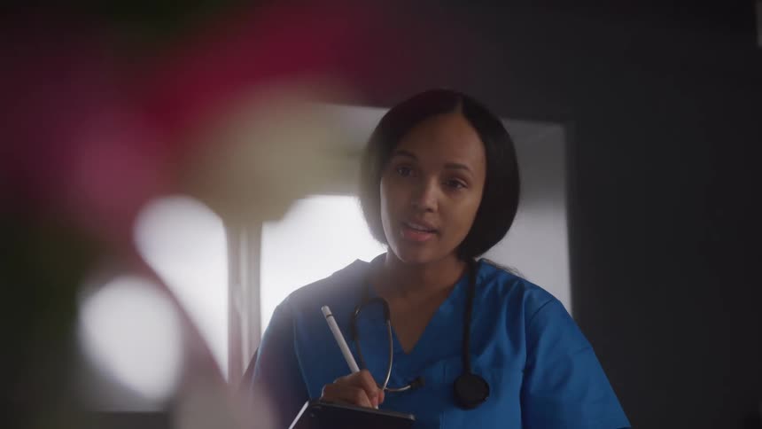 A nurse with a stethoscope around her neck is looking at a patient while holding a tablet and a stylus while wearing blue scrubs