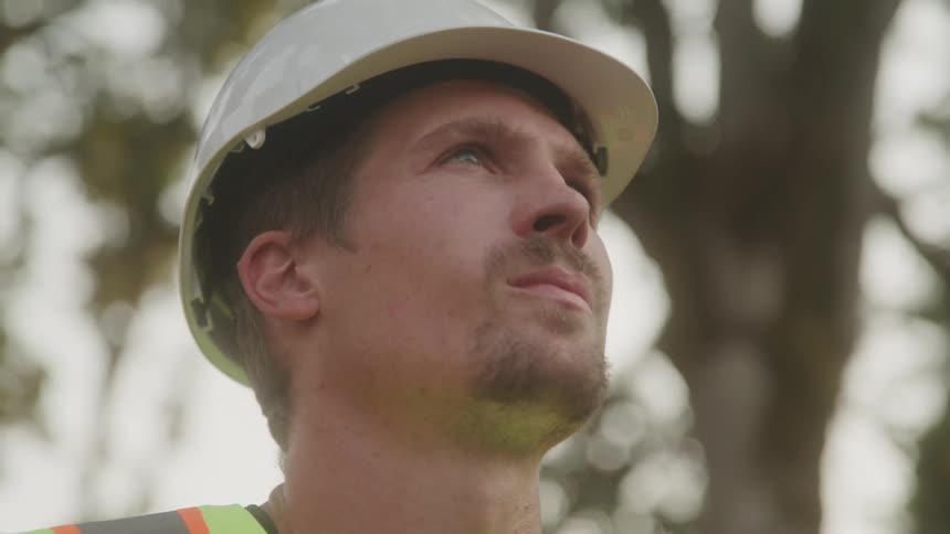 A male construction worker stands outdoors under a tree wearing a green vest and a hard hat