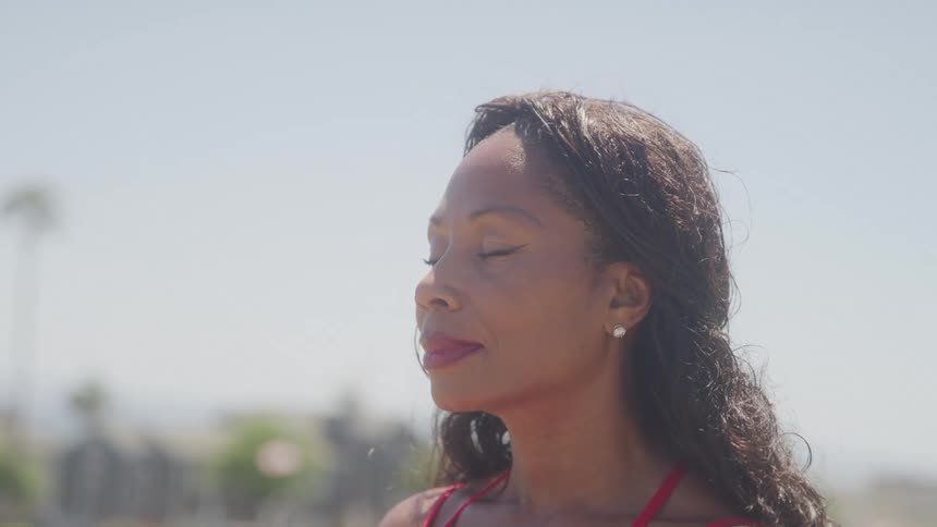 A woman in a red outfit stands outside with closed eyes basking peacefully in the midday sun