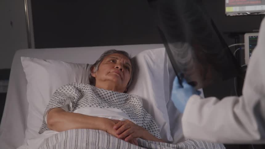 Doctor in gloves with an imaging scan talking to an elderly lady patient lying in the hospital bed