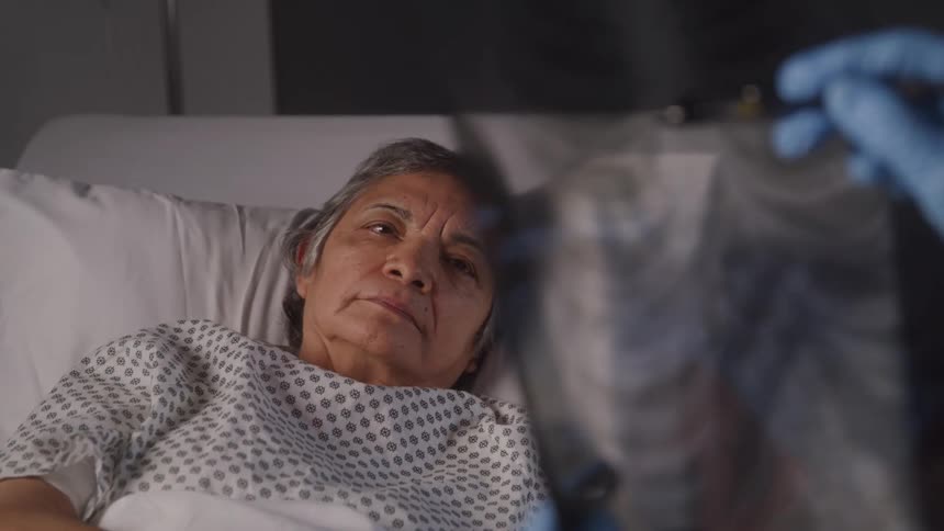 An older woman in a hospital gown lies in bed attentively observing an xray held by a gloved doctor