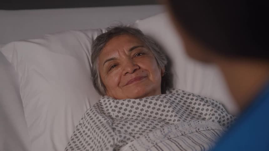 An elderly woman lies on a hospital bed with someone sitting beside her