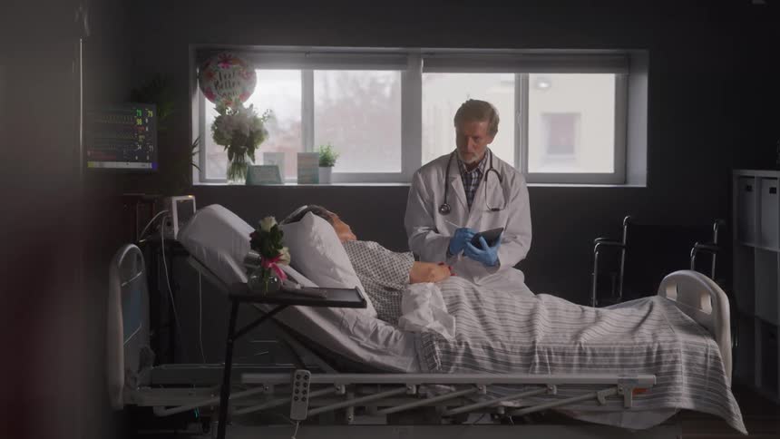A woman is sitting in a hospital bed with a doctor leaning over her holding a tablet the room has large windows with a view of a cloudy sky and trees