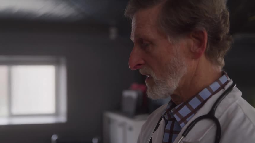 A male doctor wearing a white coat with a stethoscope hanging over his shoulder talks to a patient