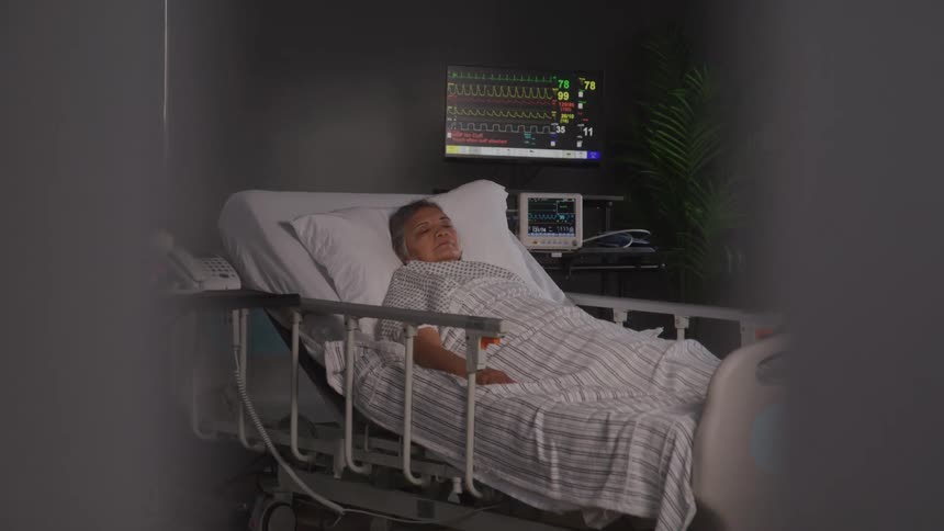 An older woman lies calmly in a hospital bed with a heart monitor displaying vital signs behind her