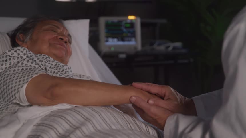An elderly patient lies in a hospital bed holding hands with a visitor