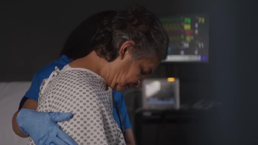 A young female medical personnel consoles an elderly female patient sitting on a bed in a hospital room undergoing treatment