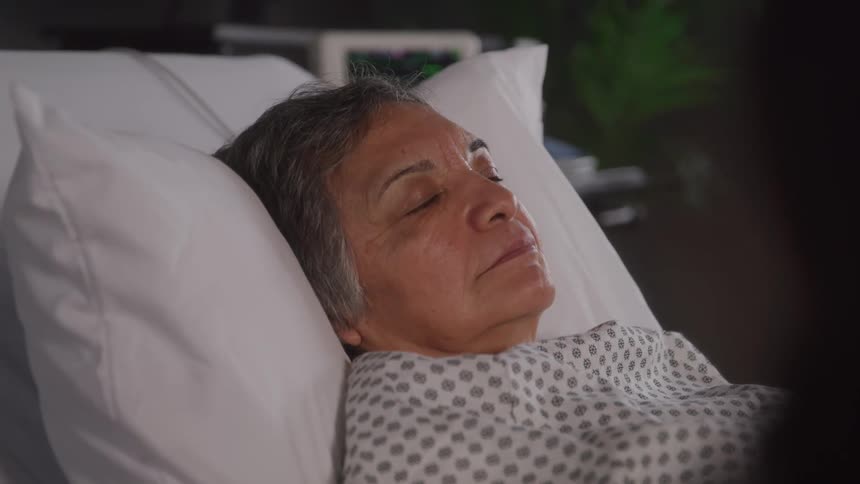 An older woman lies in a hospital bed with a somber expression looking sad or worried as she rests on a pillow