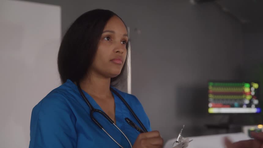 A woman in blue scrubs with a stethoscope around her neck reviews a chartwith a person beside a hospital bed
