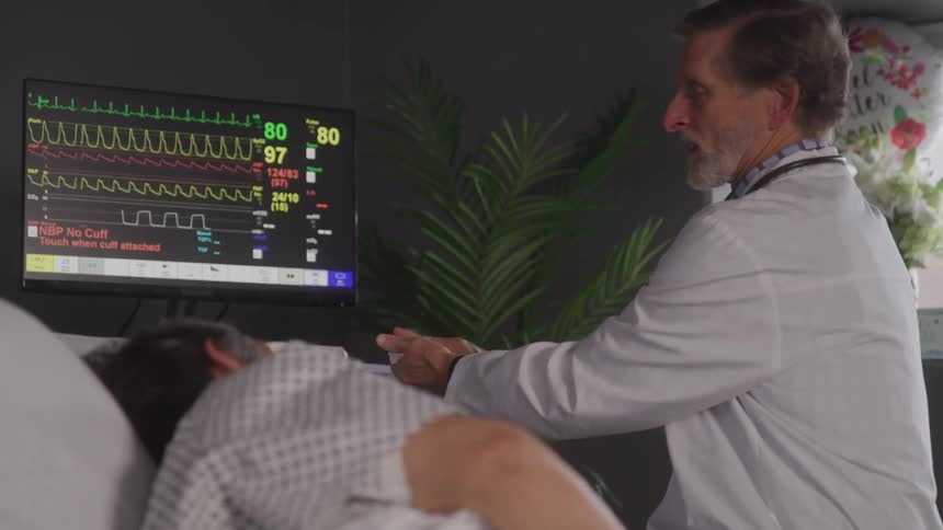 The video shows a male doctor sitting at the bedside of an elderly patient who is lying in a hospital bed the patient is looking towards the doctor and a heart rate monitor is visible in the background