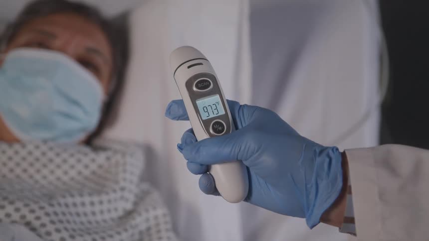 A healthcare worker in blue gloves is holding a digital thermometer with a reading of 973 degrees a person wearing a surgical mask is partially visible and out of focus in a hospital bed in the background