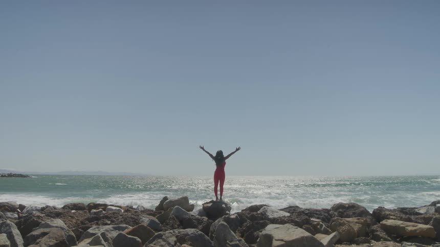 The video shows a woman standing with her back to the camera with the ocean and rocks in front of her she is wearing a red outfit and has her arms stretched out to the side