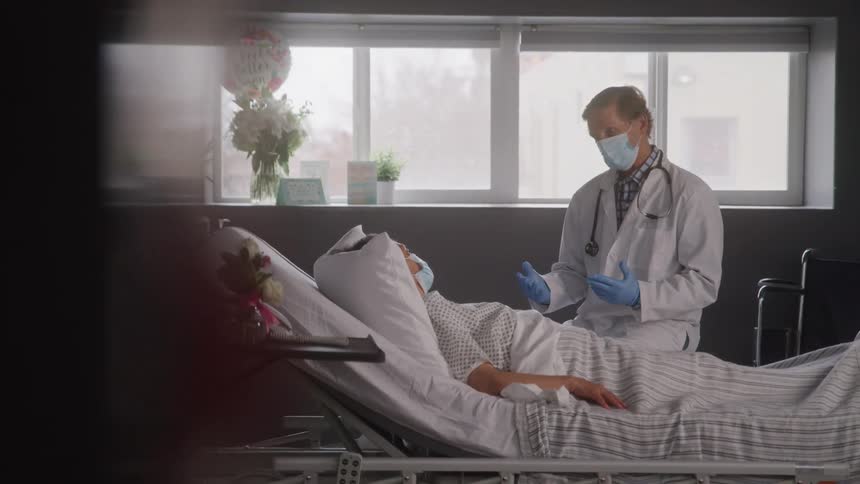 As the camera slowly reveals the scene a doctor wearing a mask and a patient both look at each other in a hospital room with a get well soon balloon visible in the background