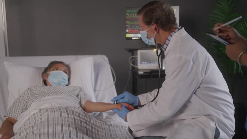 The video shows a male doctor wearing a mask and gloves using a stethoscope to check the pulse of a patient lying in a hospital bed the patient is also wearing a mask and is looking at the doctor