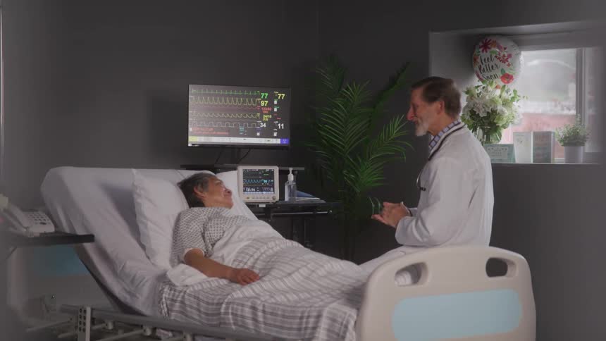 A doctor wearing a white coat and a stethoscope sits on a hospital bed and speaks to a patient who is lying in the bed
