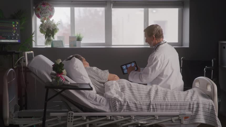 The video shows a doctor in a white coat speaking with an elderly patient who is lying in a hospital bed the doctor is holding a tablet and gesturing towards it while the patient looks up at them