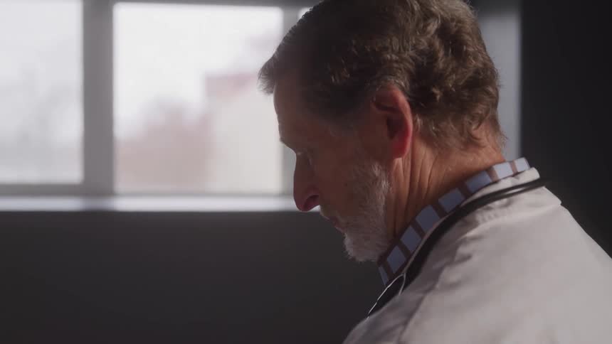 A male doctor with graying hair and a beard is shown in a closeup side profile shot he is wearing a stethoscope and appears to be looking down with a serious expression