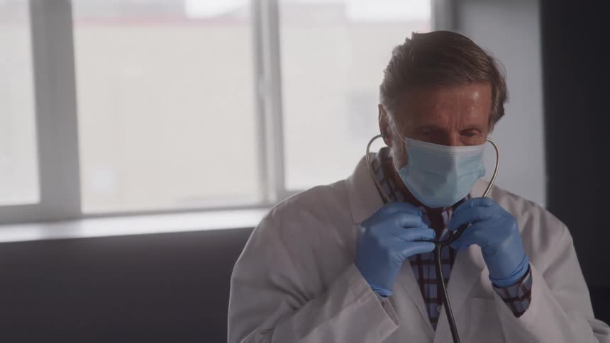 A doctor wearing a face mask a white coat and blue gloves holds a stethoscope to his ears while looking down