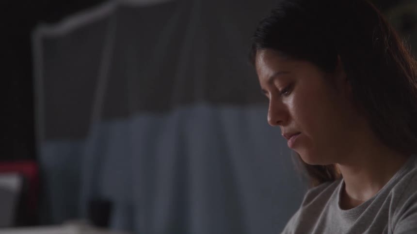 A woman with long dark hair wears a grey shirt and stares down at an object outside of the frame with a serious expression and then looks up