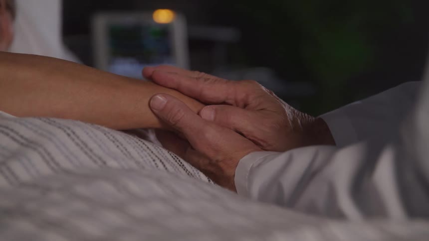 Elderly person holds the hand of a patient lying in a hospital bed