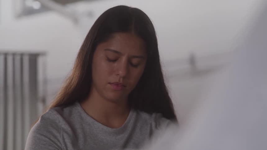 A closeup of a womans face shows her looking down with a somber expression with a soft blurred background and light coming from behind her