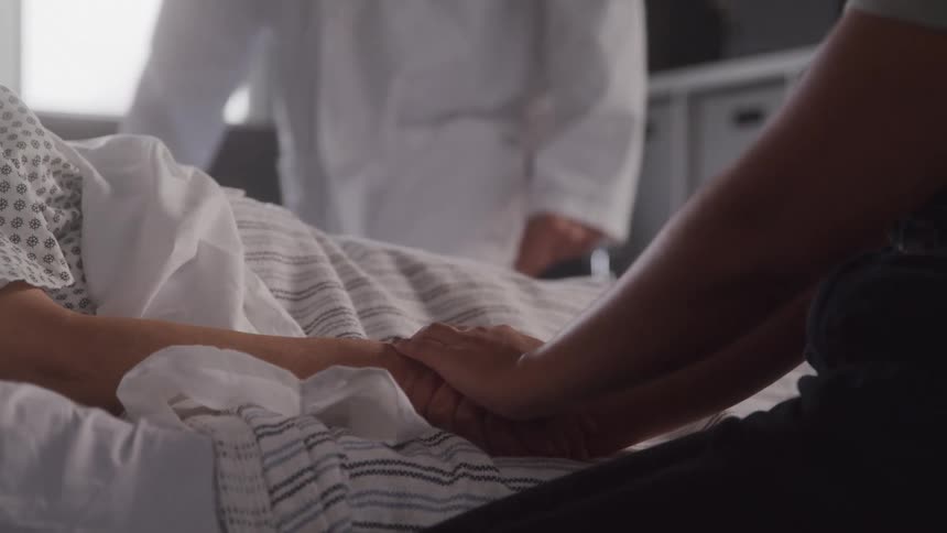 A person holds a patients hand as the patient rests in a hospital bed while a doctor in a lab coat sits behind them