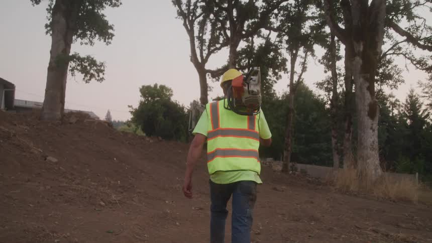 A worker in a highvisibility vest and hardhat walks across a dirt mound at a construction or landclearing site carrying a piece of heavy equipment on his shoulder