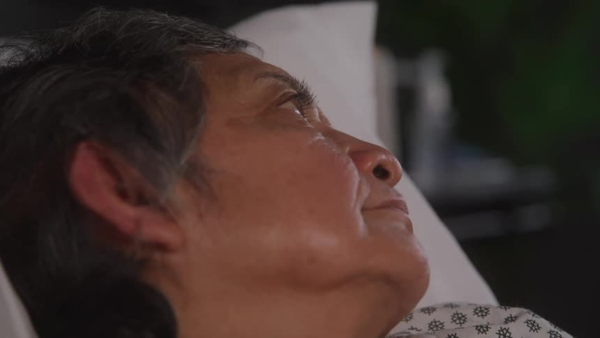 A closeup shot of an older woman lying in a hospital bed looking up and to the side with a serious or pensive expression