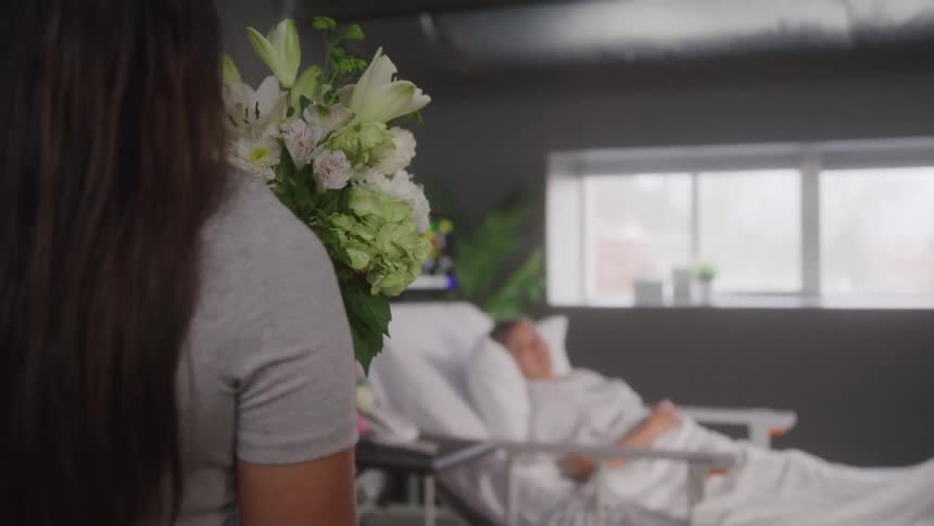A young woman carrying a flower bouquet approaches an elderly sick female patient lying in a hospital room during daytime