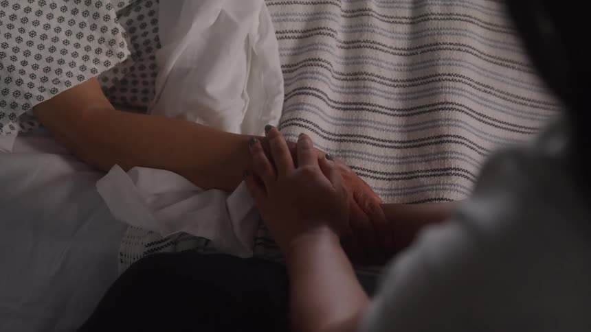 A woman gently touches the hand of a patient who is lying in a hospital bed with one arm resting on the blankets