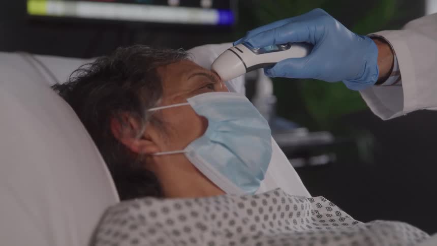 Close up of a medical personnel checking an elderly female patients temperature with a device while she is lying down in a hospital bed