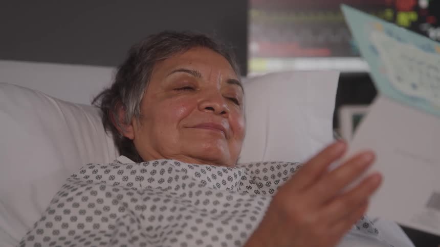 Older female patient laying in a hospital bed reading a get well card