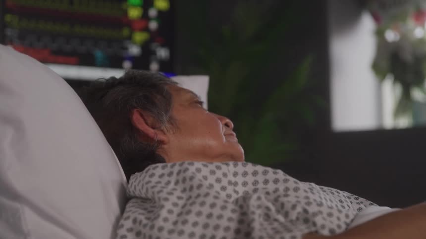 The video shows an overhead shot of a person lying in a hospital bed facing away from the camera a medical monitor with a glowing screen is visible in the background