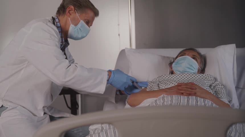 A medical professional wearing a white coat a stethoscope and a face mask injects a patient who is lying in a hospital bed while also wearing a face mask