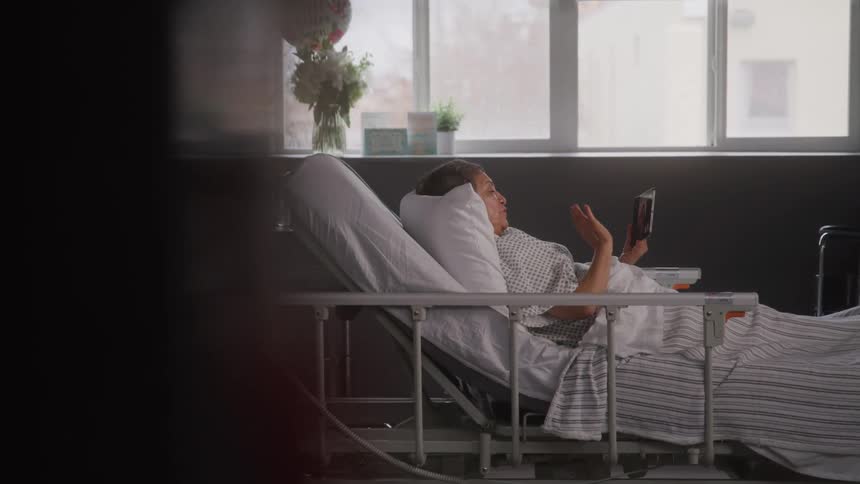 An elderly female patient waves over a video call with a tablet device while lying on a bed in a hospital room during daytime