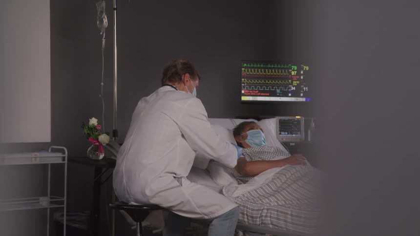 The video shows a healthcare professional in a white coat tending to a patient who is lying in a hospital bed the patient is wearing a face mask and a medical monitor with various vital signs is visible in the background