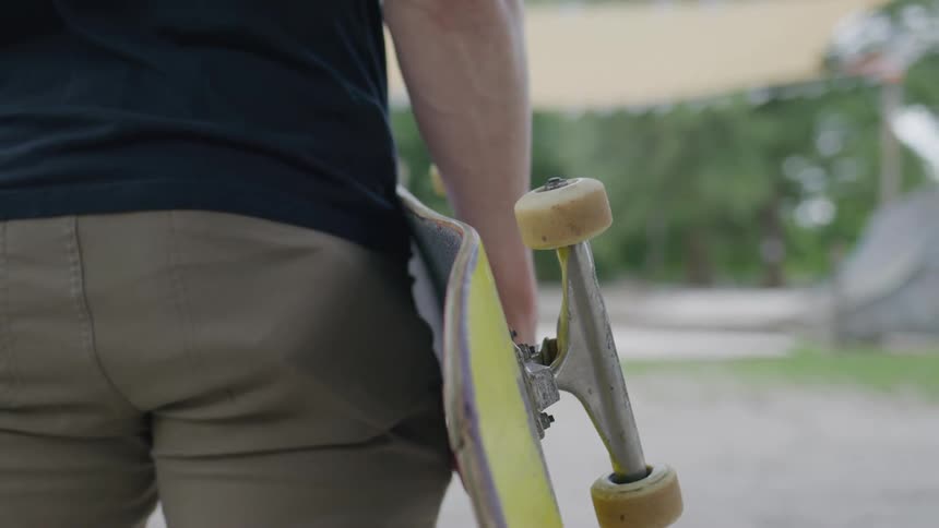 A closeup shot of a man from the waist down holding a skateboard he is wearing a dark tshirt and khakicolored pants