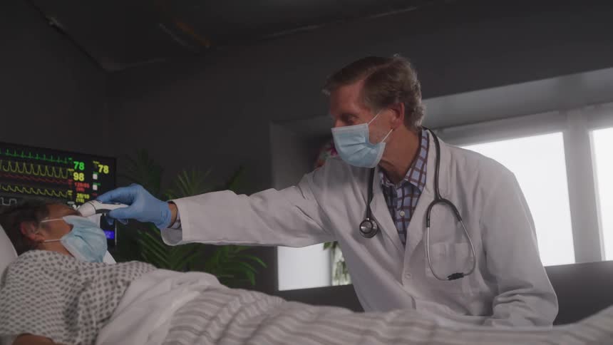 A male doctor wearing a white coat and a face mask measures the temperature of a patient in a hospital bed with a thermometer