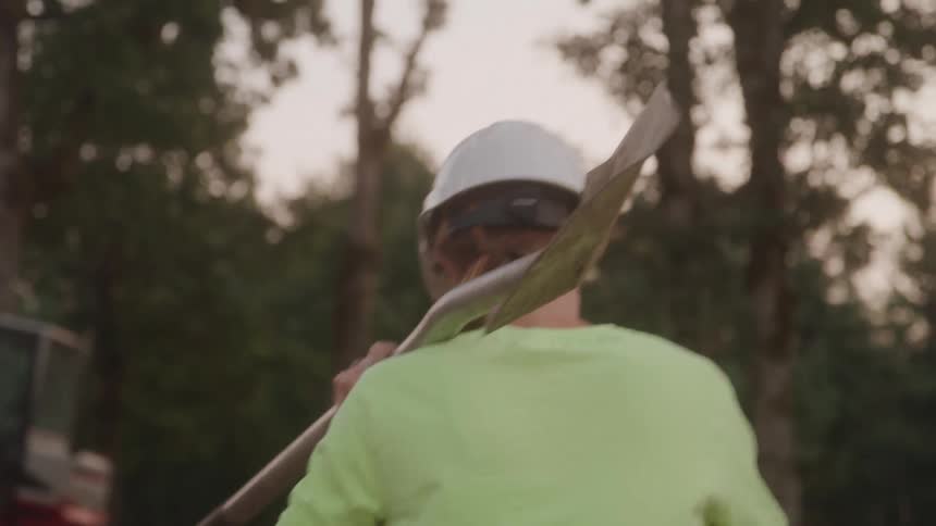 Person in a green shirt and cap carries a shovel resting on the shoulder