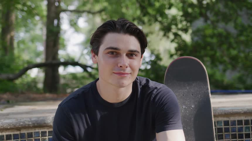 A young man sitting in a public park at midday with a skateboard resting beside him