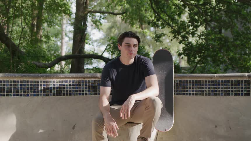 The video shows a young man sitting on the edge of a concrete structure in a skatepark he is wearing a black shirt and beige pants and a skateboard is propped up next to him