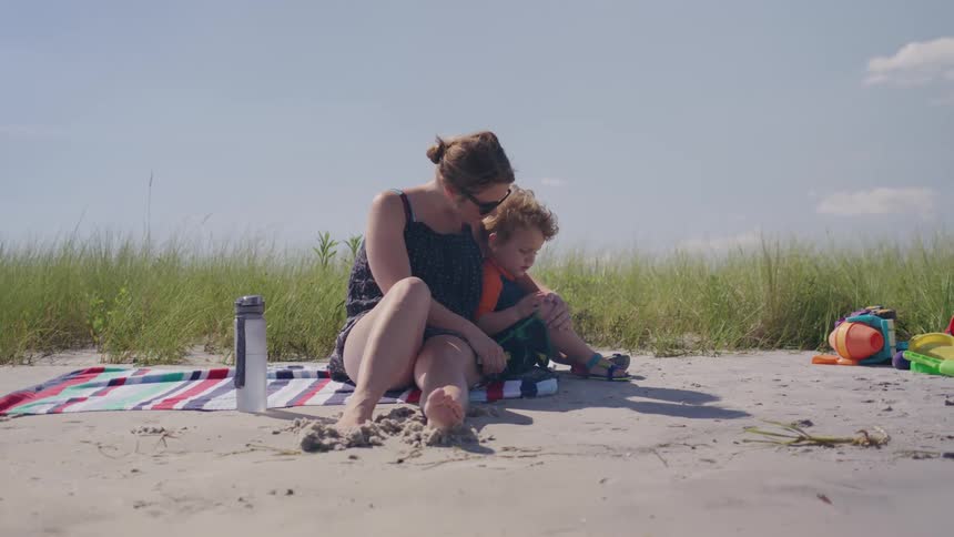 The video shows a mother and her young child sitting together on a sandy beach they are surrounded by tall grass and various beach toys are scattered nearby