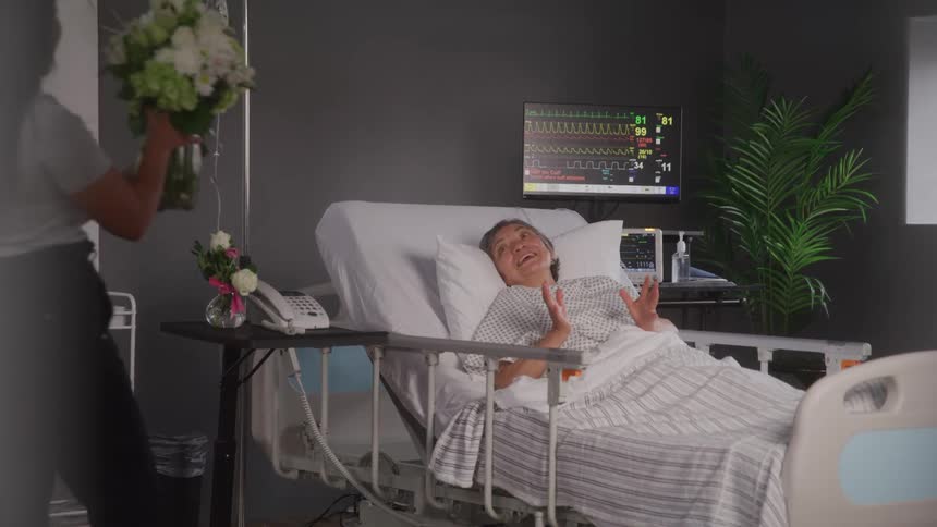 A young woman visits an elderly woman lying in a hospital bed looking excited as she brings flowers to her at midday