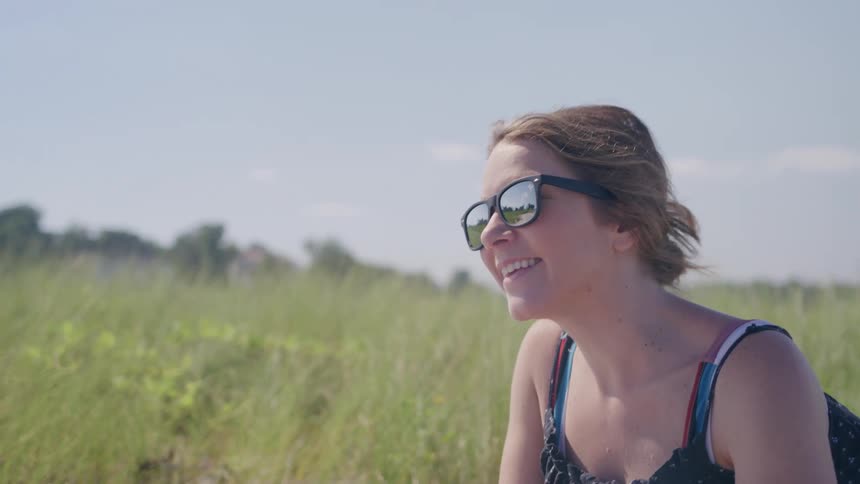 A woman wearing sunglasses and a patterned tank top is smiling and looking at a person just out of frame she is sitting outside in a field of tall grass on a sunny day