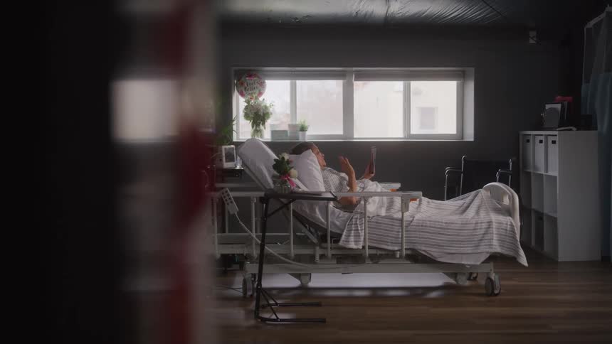 A female patient in a hospital bed raises her arms waving goodbye as she lies in a room with a window medical equipment and a gift bouquet