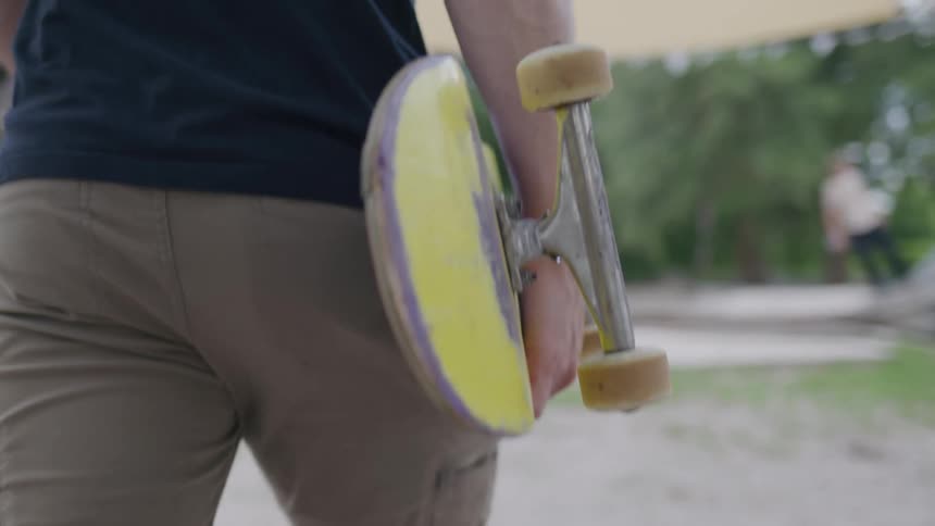 A man walks with his back to the camera carrying a skateboard with a yellow underside and tan wheels