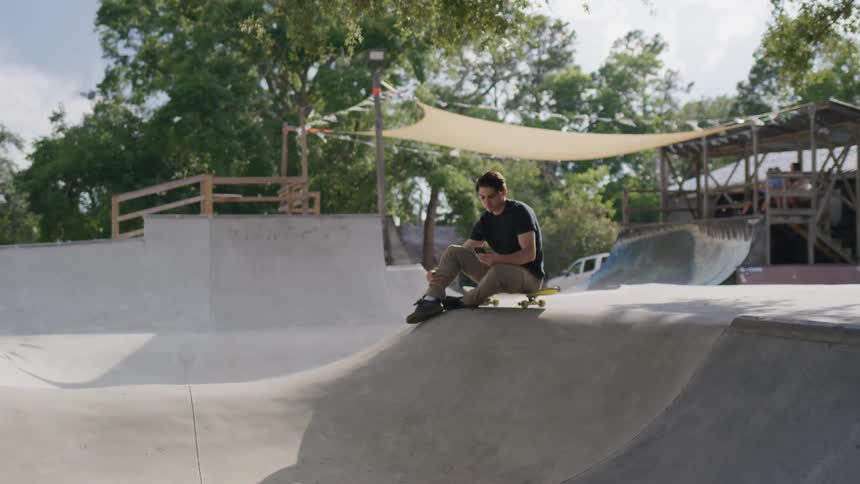 The video shows a young man sitting on his skateboard at the top of a ramp in a skate park he is wearing a black tshirt and jeans and is looking at his phone with a serious expression